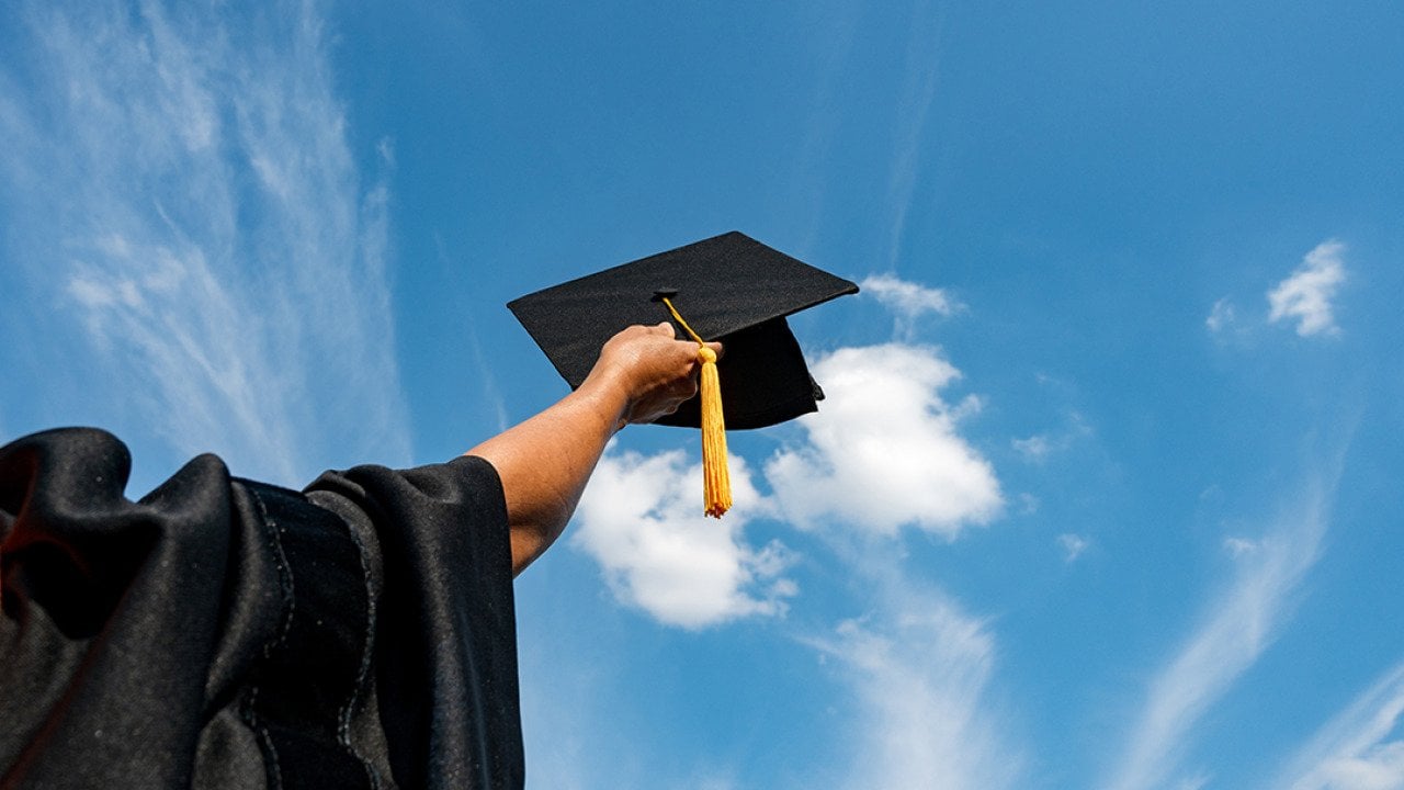 A graduate student holding up a graduation cap in the sky.