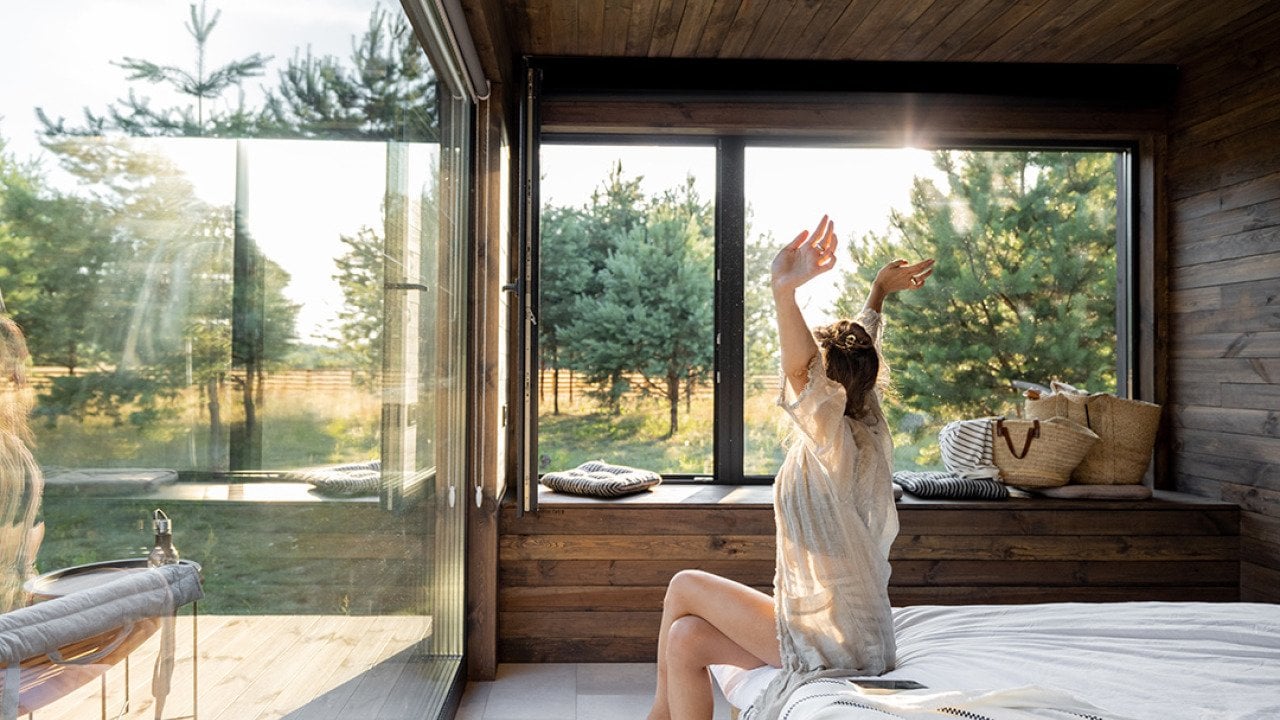 Woman stretching as she is waking up in a modern country house with glass windows.