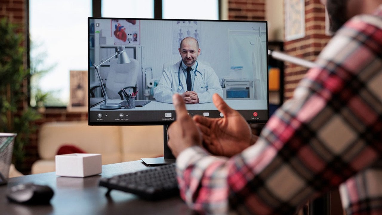 Computer screen showing telehealth call with male balding doctor sitting in office with stethoscope around his neck, while person dressed in plaid long sleeve shirt in foreground converses on video call with his back to the camera.