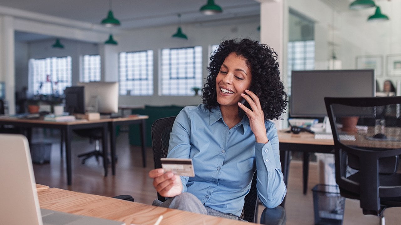 Businesswoman in her workplace looking at her credit card while on the phone.
