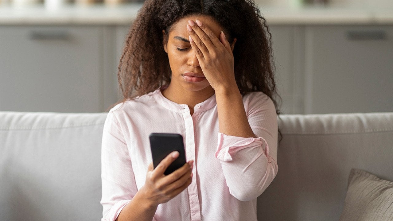 A photo capturing the tension of a young African American woman sitting on her couch, visibly overwhelmed as she looks at her smartphone in a neatly arranged living room.