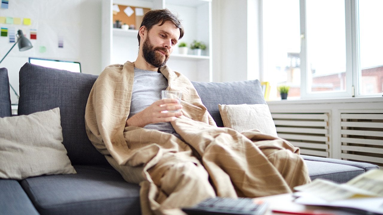 Sick man wrapped into blanket sitting on sofa in front of table with papers while staying at home.