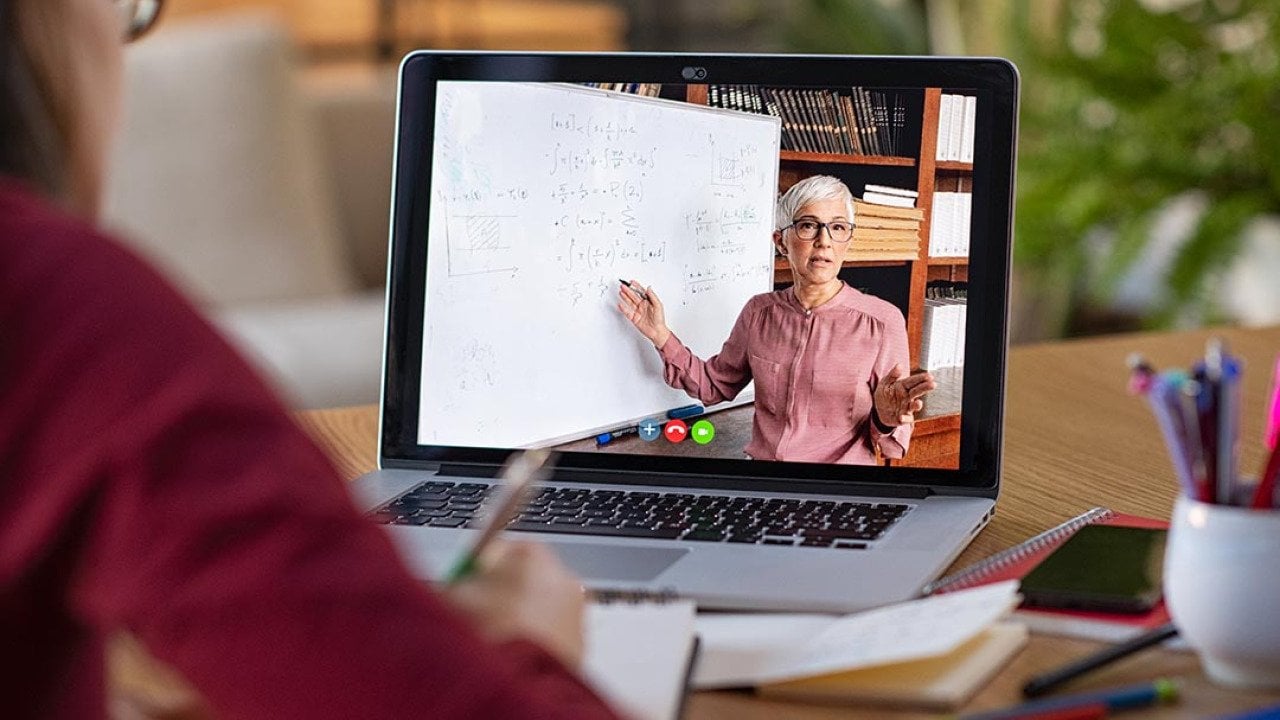 A video on a computer screen video showing an older teacher demonstrating lesson on whiteboard, with student watching in foreground.
