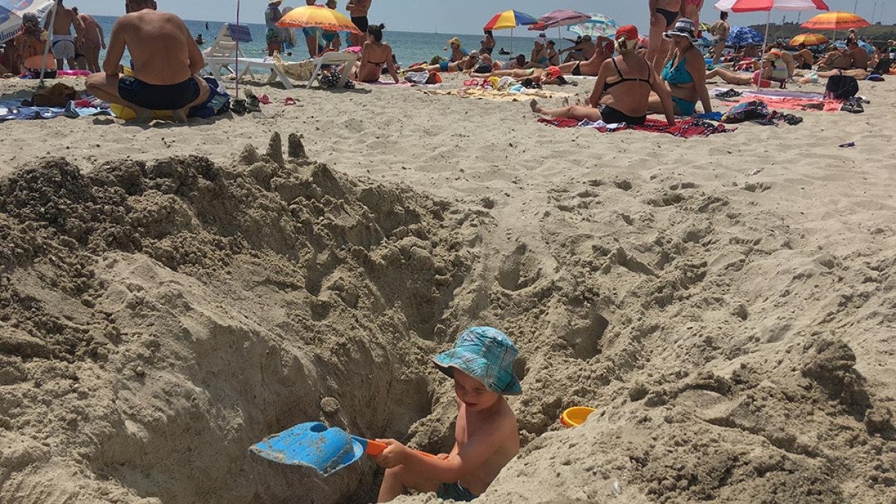 Child wearing a blue hat digging a giant hole in sand at the beach that is higher than him on both sides.