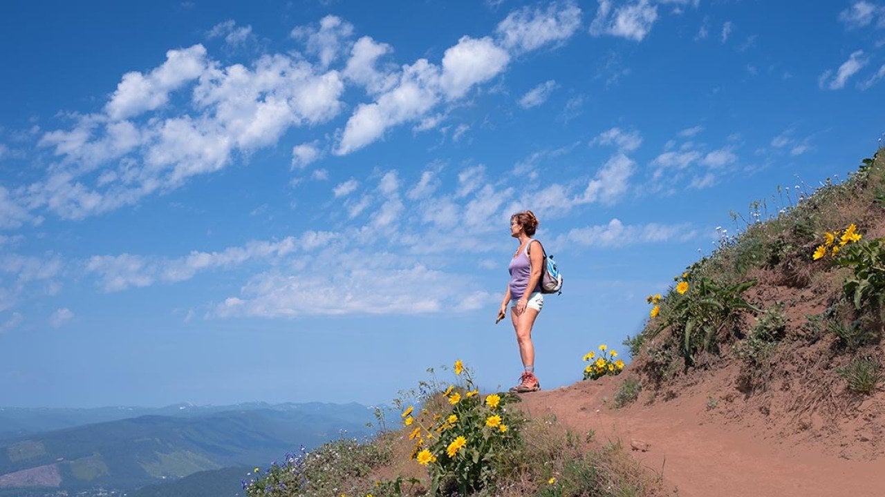 Woman looks out toward view on dirt mountain trail with yellow flowers, hiking in summer with sky and clouds in background near Portland, Oregon.