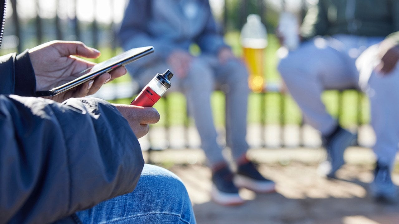 Teenager holding a vape while scrolling on the phone at the park.