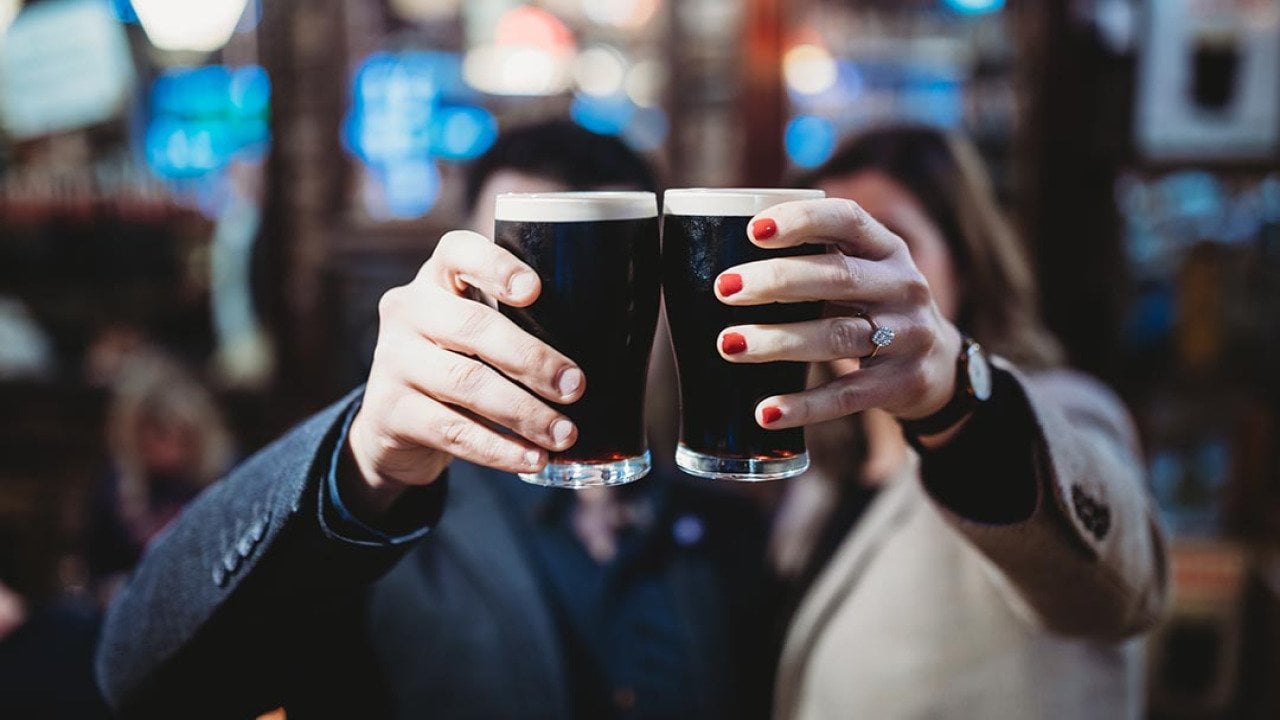 Two people holding up a pint each of stout beers to cheers, which obscures their faces from the camera in a bar.