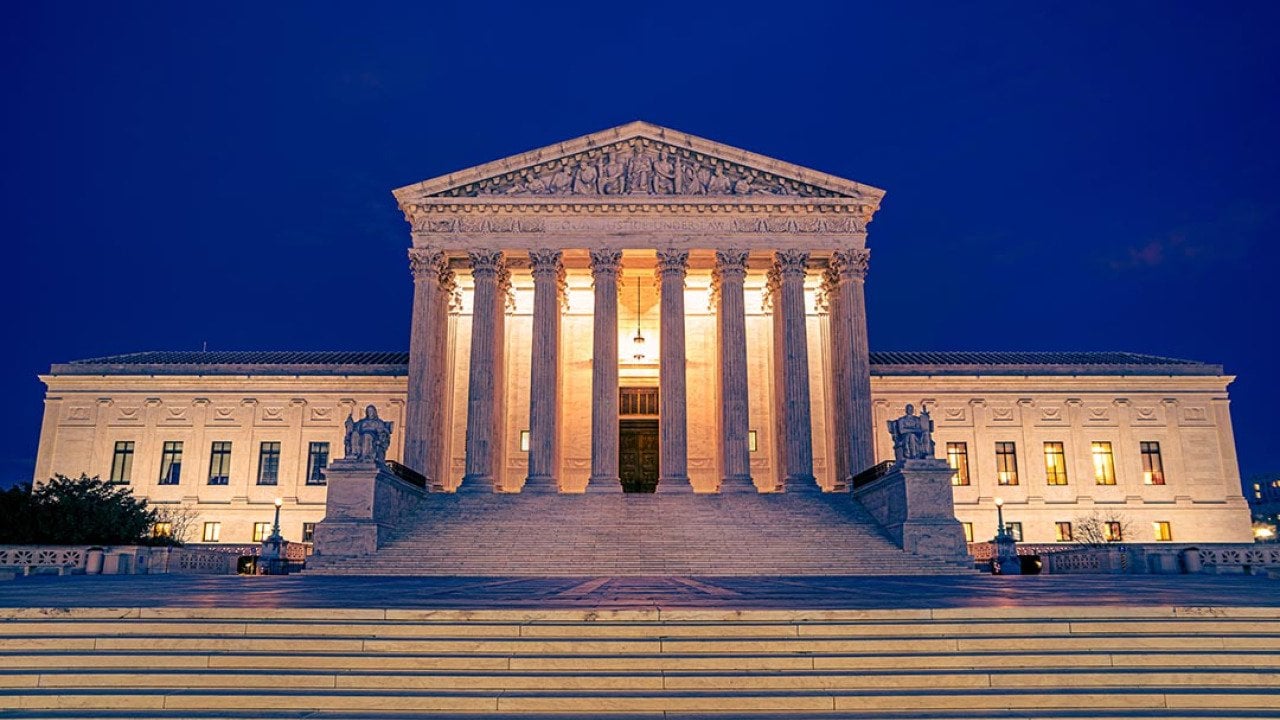 United States Supreme Courts building illuminated at night in Washington DC with steps and plaza in foreground.