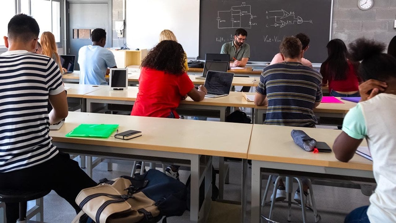 View from back of classroom of multiracial high school students in class using laptops while teacher marks exams.