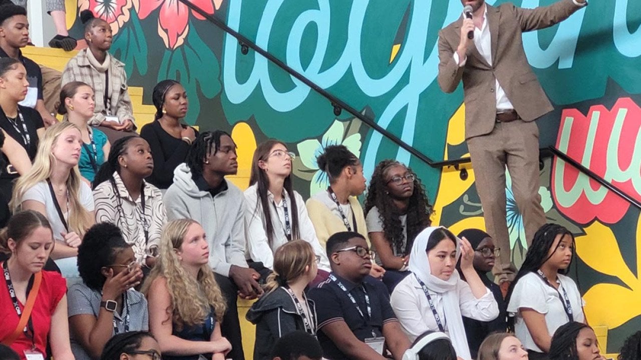 Side view of rows of high school students sitting on bleachers listening to a speaker wearing a tan suit talking into a mic.