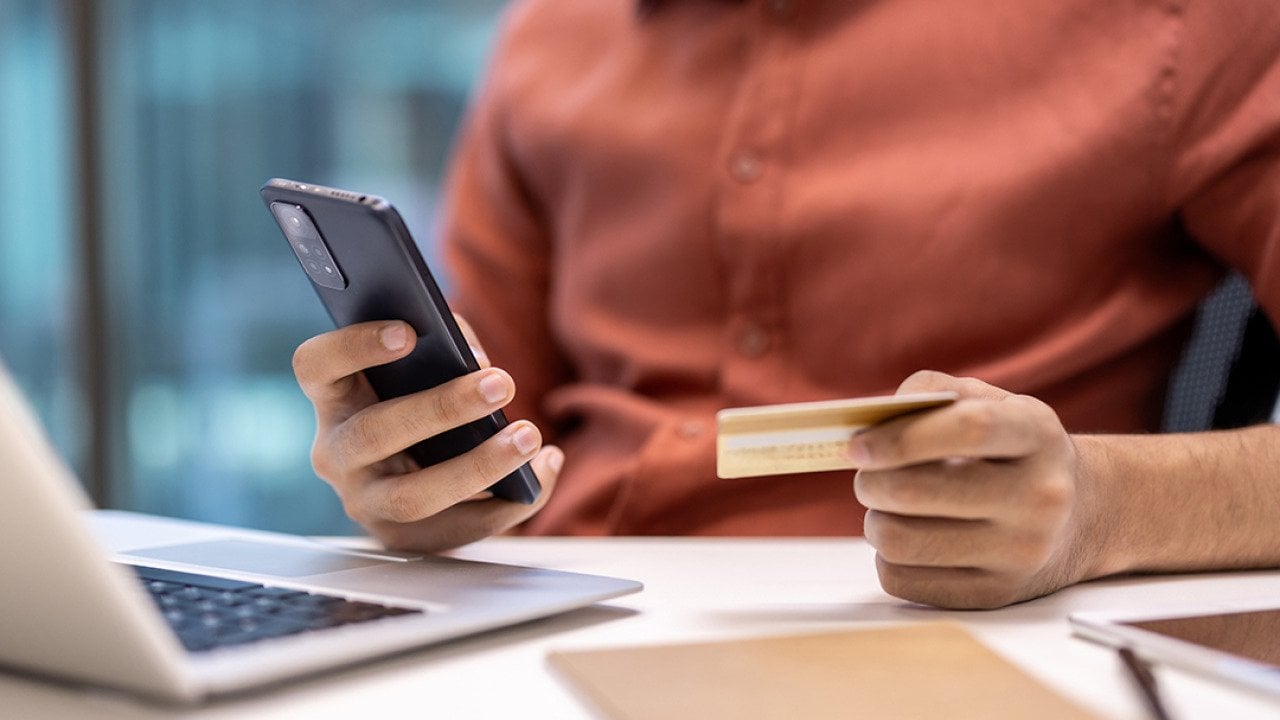 Businessperson holding a credit card while using a phone and laptop at an office desk.