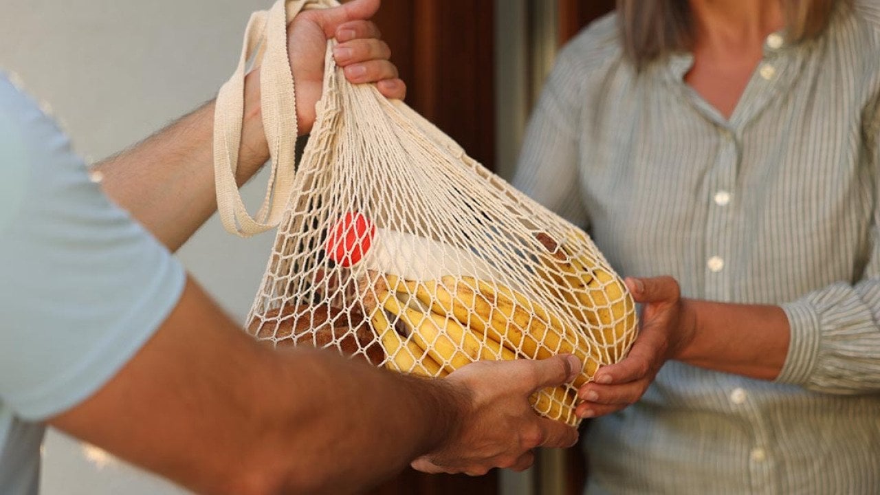  Man hands a net bag of groceries to woman in the doorway, their faces are not visible.
