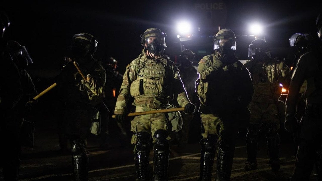 Nighttime view of a line of federal immigration agents in riot gear and face shields facing the camera near Glass House Farms facility in Camarillo, with police spotlights in background.