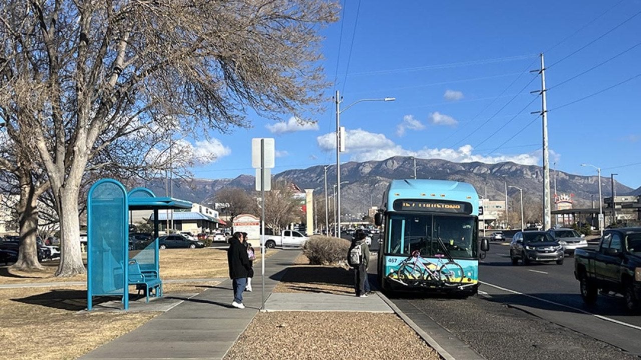 Bus riders board a blue bus in Albuquerque, next to a turquoise metal bus stop and with mountains and cloud in distance.
