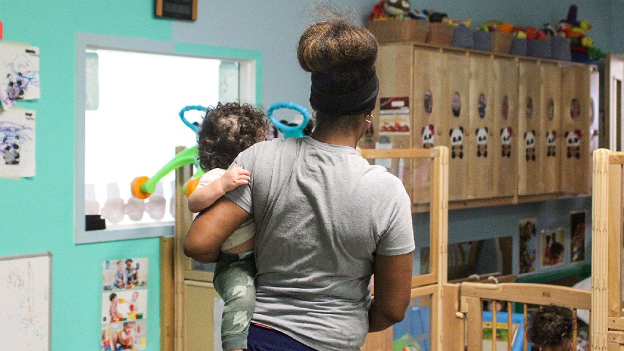 A child care teacher carrying an infant in a child care room.