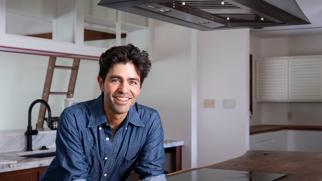 Adrian Grenier stands beneath his island-mounted CopperSmith® range hood, featuring a smooth bronze finish, paired with a handcrafted wood countertop for a warm, natural aesthetic.