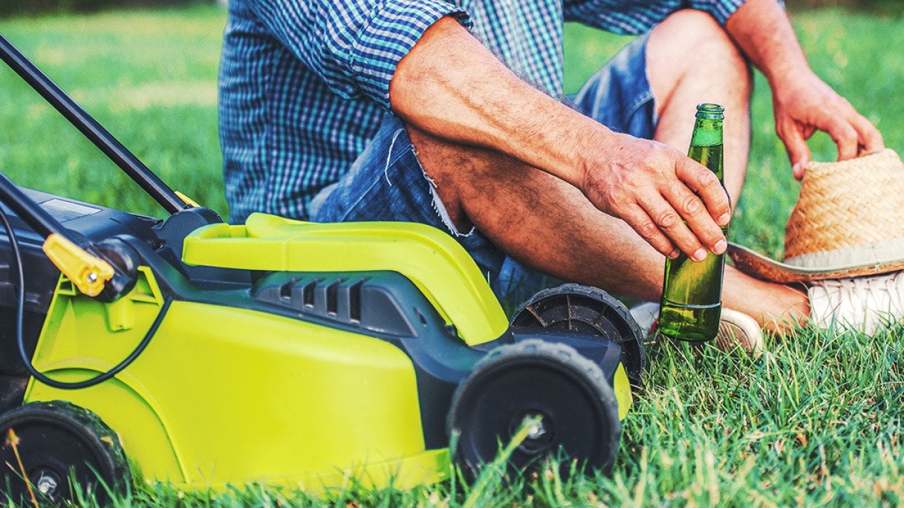 Senior man enjoying a bottle of beer beside a mower in his lawn.
