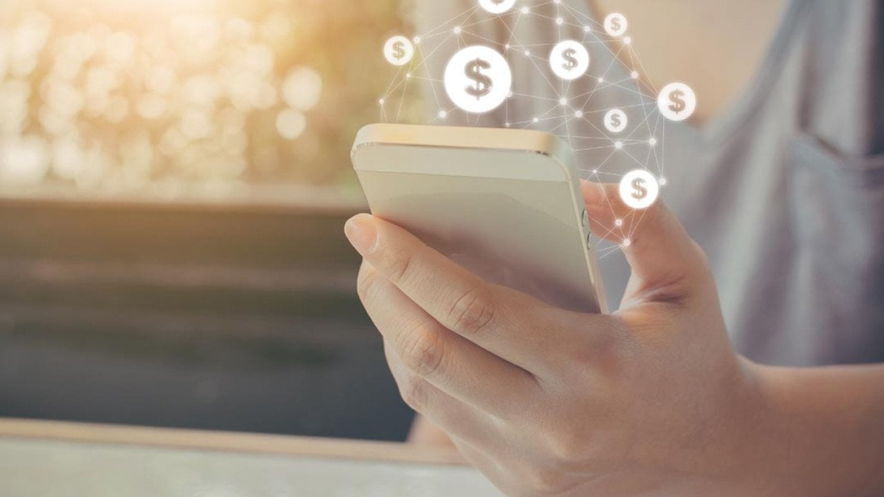 Hand holding mobile phone with dollar sign icons overlaid, symbolizing ecommerce transactions made on smartphones.