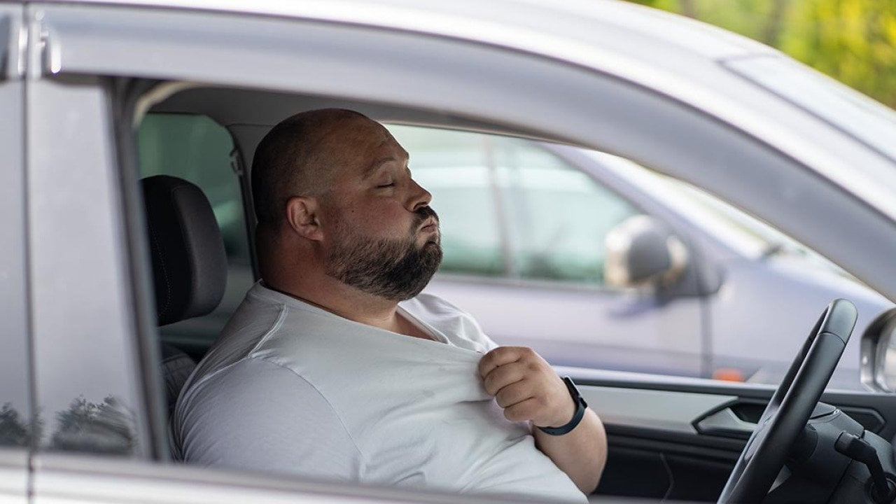 A bald driver with closed eyes seen through the passenger side window pulls at the collar of a white t-shirt and exhales.