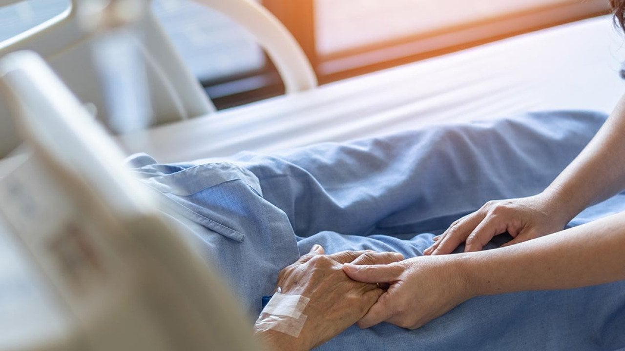 Elderly senior patient in hospital bed, whose face and torse are not visible, has hand held by younger caretaker.