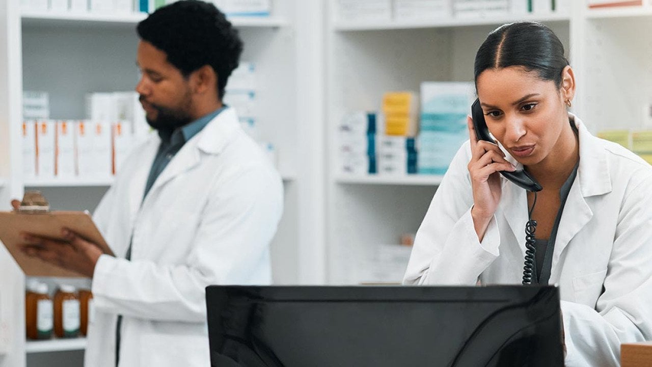 Two pharmacy employees work behind the counter in a pharmacy, one on the phone and looking at a computer and the other writes on a clipboard.
