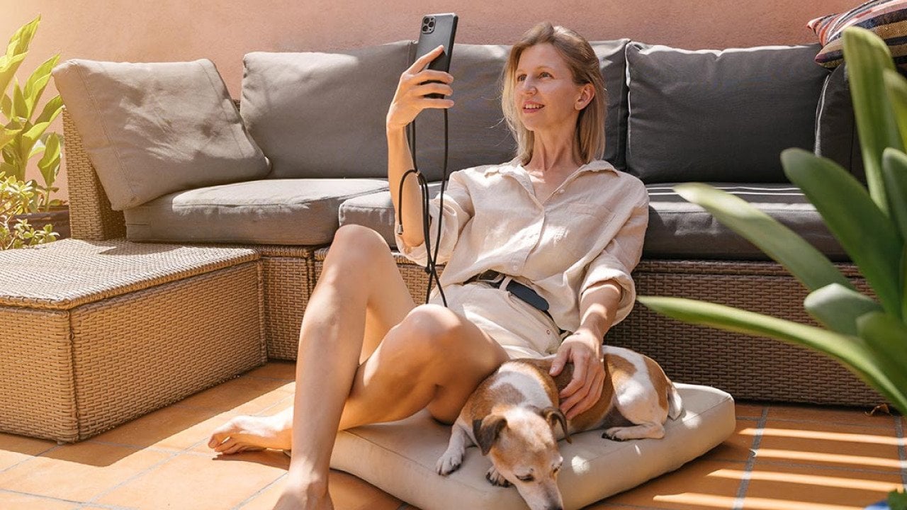 Woman wearing neutral button up top and shorts sitting on a floor cushion leaning against a couch, next to her dog, facetiming on her phone.