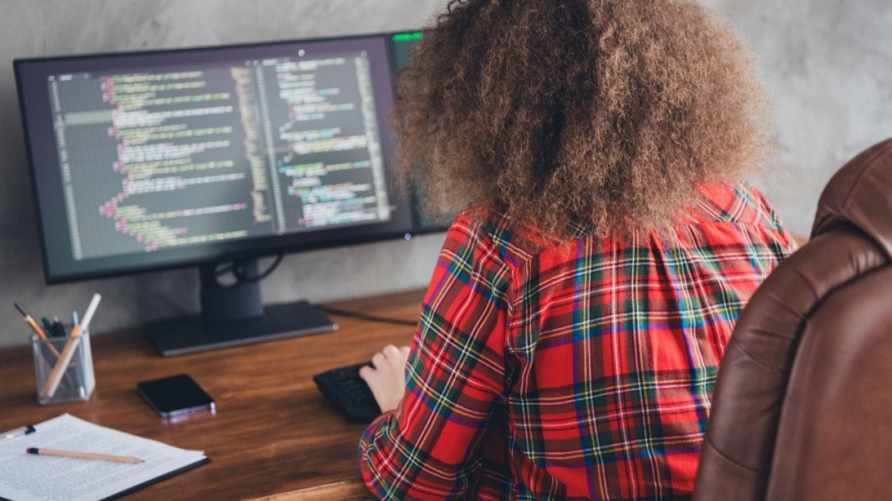 Rear view of a woman with curly hair working at a computer desk displaying code in a casual office setup wearing a checked shirt.