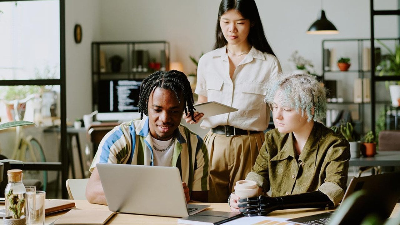 Three diverse young adults gathered around a laptop in a modern office setting.