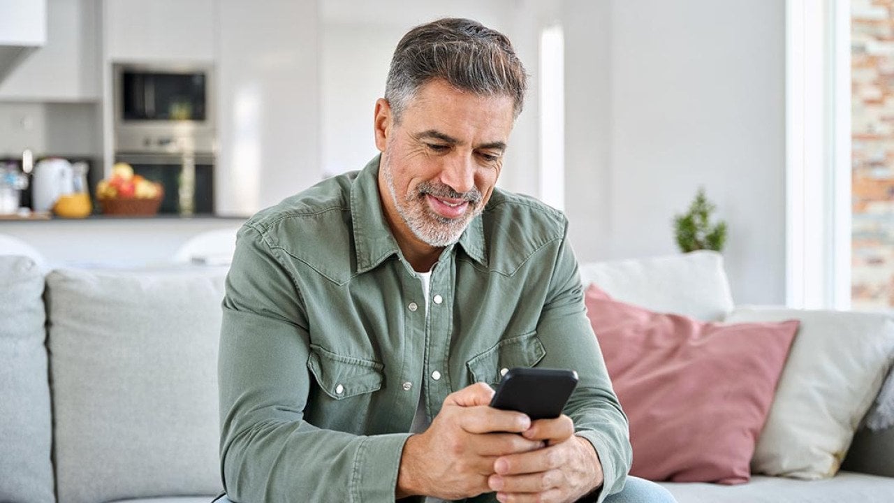 Middle aged old man using smartphone sitting on a couch with kitchen in background.
