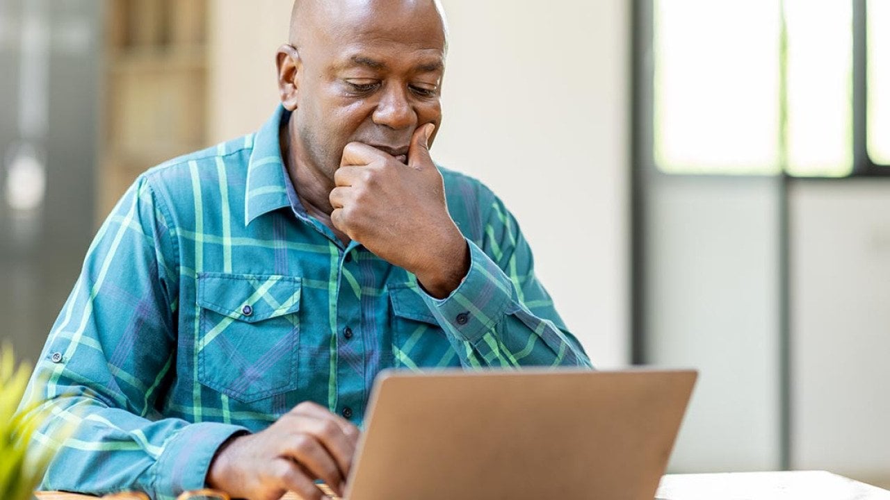 Black bald man wearing teal button up shirt using laptop computer and thinking about question in office.