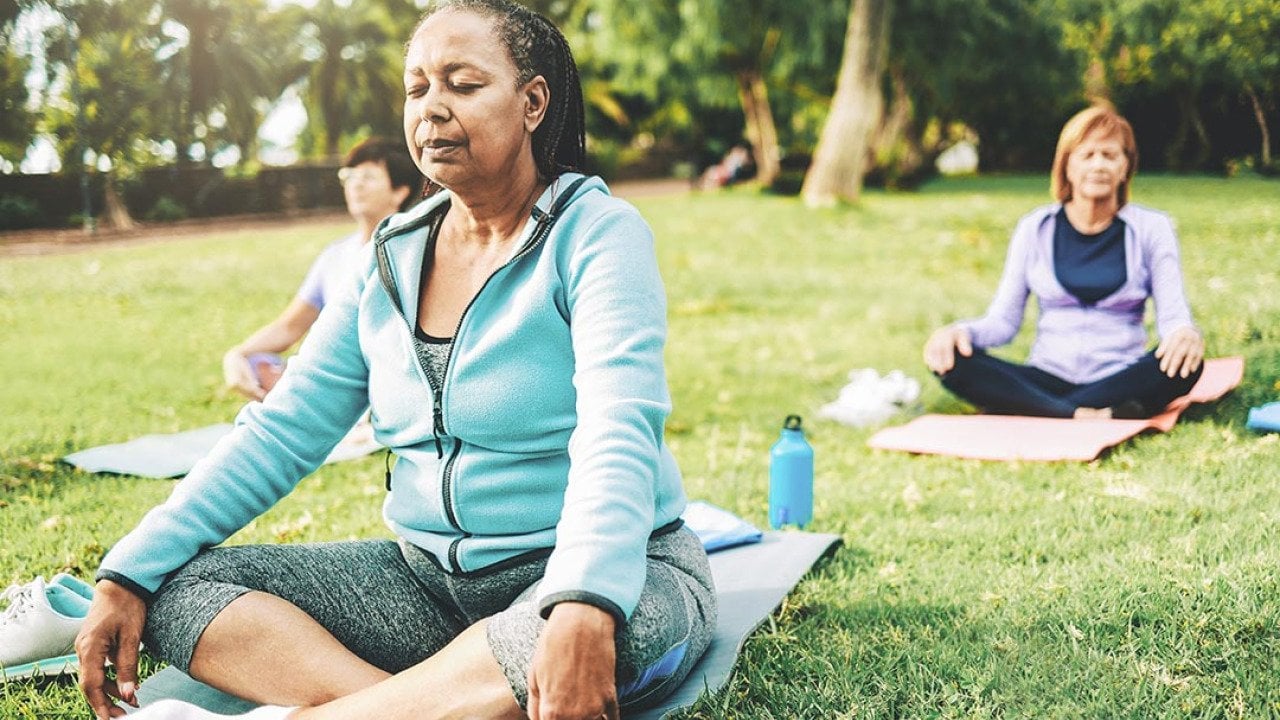 Three people in seated yoga poses on individual yoga mats on a green lawn, not sitting close together.