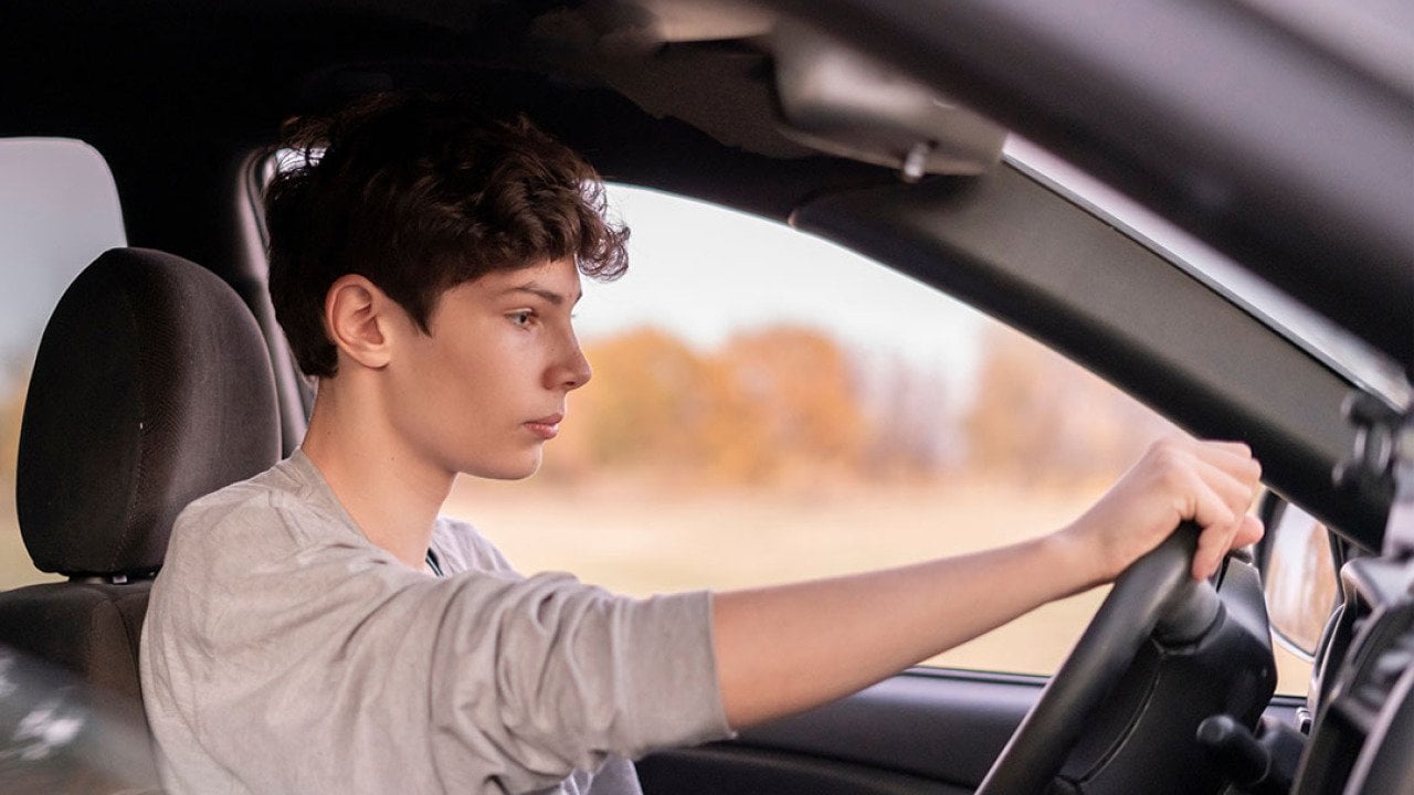 Young male driver holding steering wheel in car seen through the passenger side window.