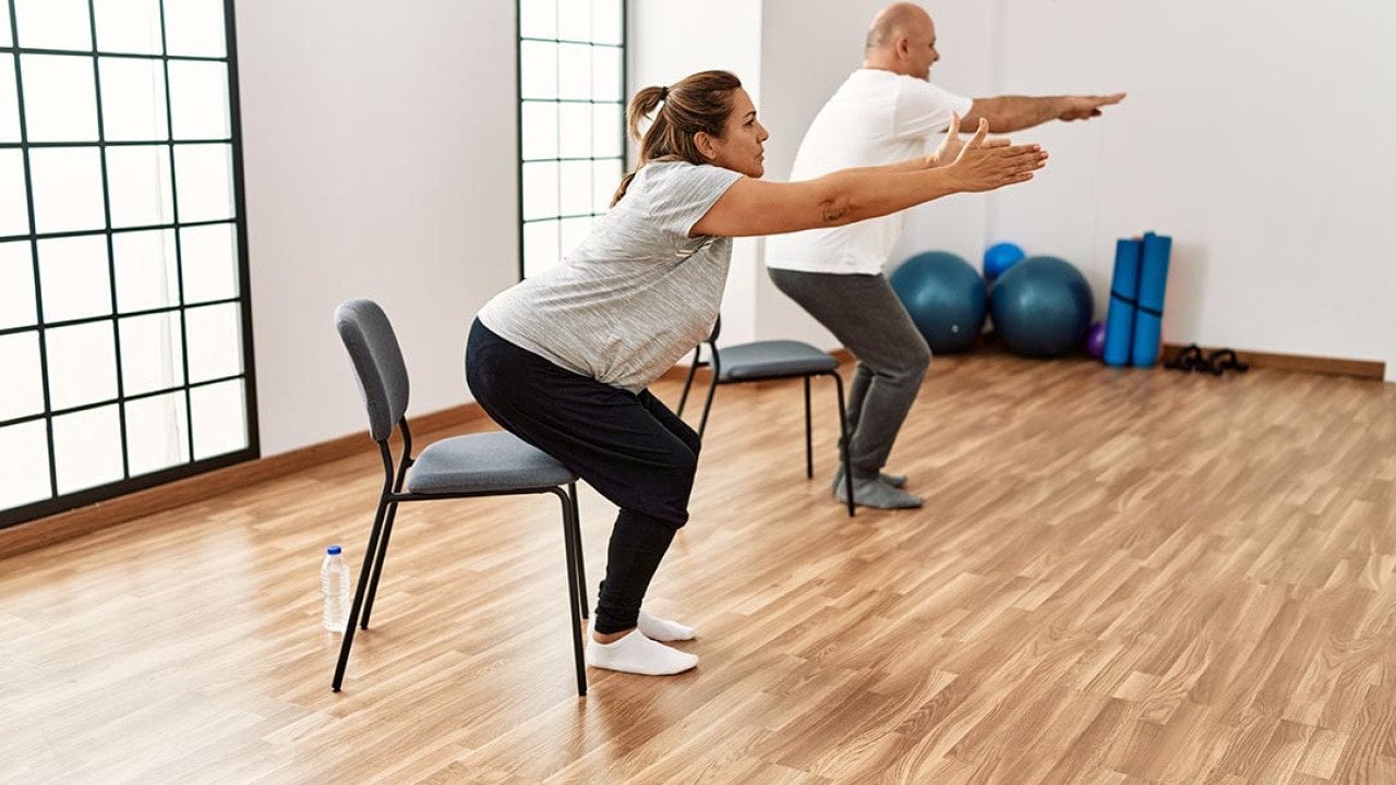Two people each squatting toward a chair with their arms out in front of them, in a yoga studio type room.
