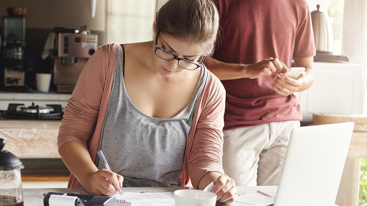 Woman doing calculations at kitchen table with laptop open and pen and paper while man with face not visible in the background is on his phone.