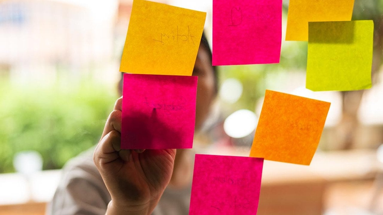 7 Pink, orange and yellow post-it notes stuck on glass with person whose face is obscured, writing on them from the other side.