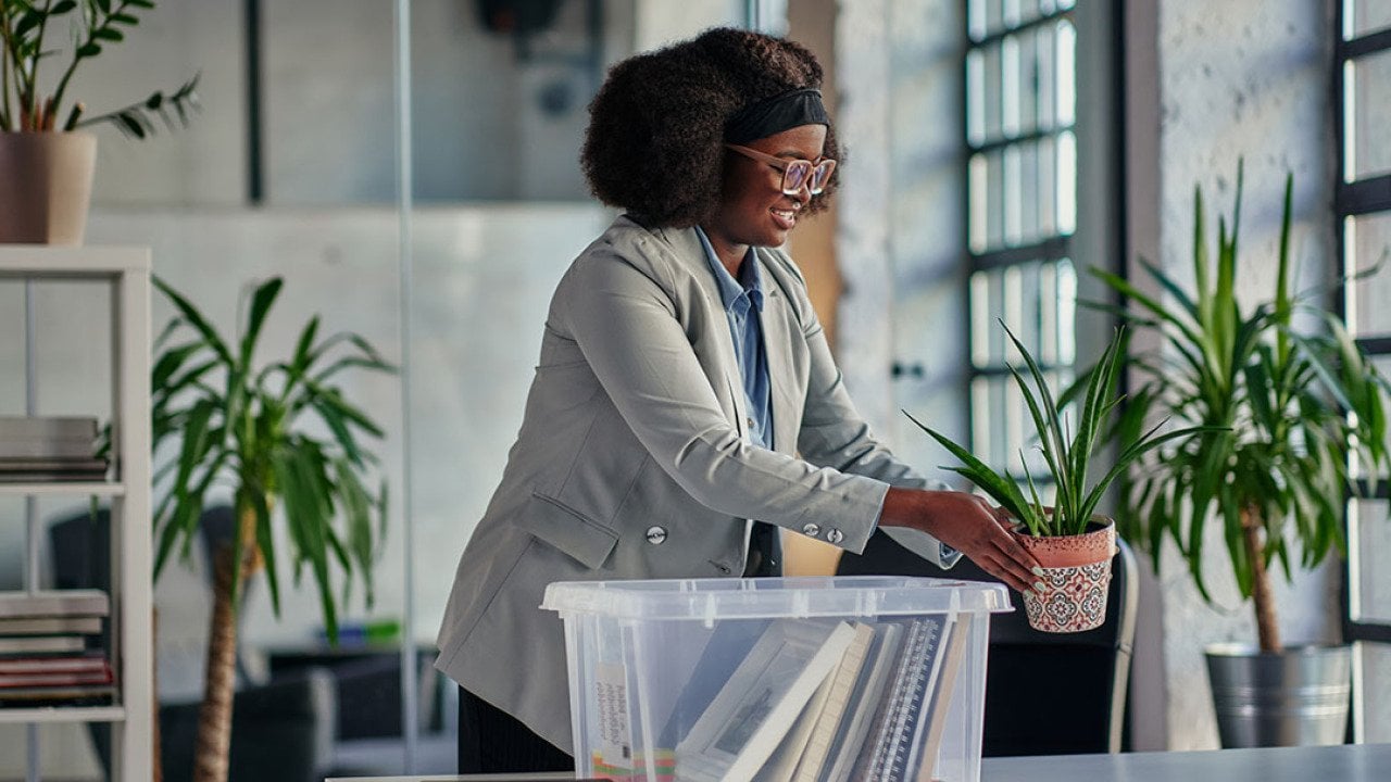 Black woman with glasses placing plants on an office desk with a clear plastic bin full of belongings on table.