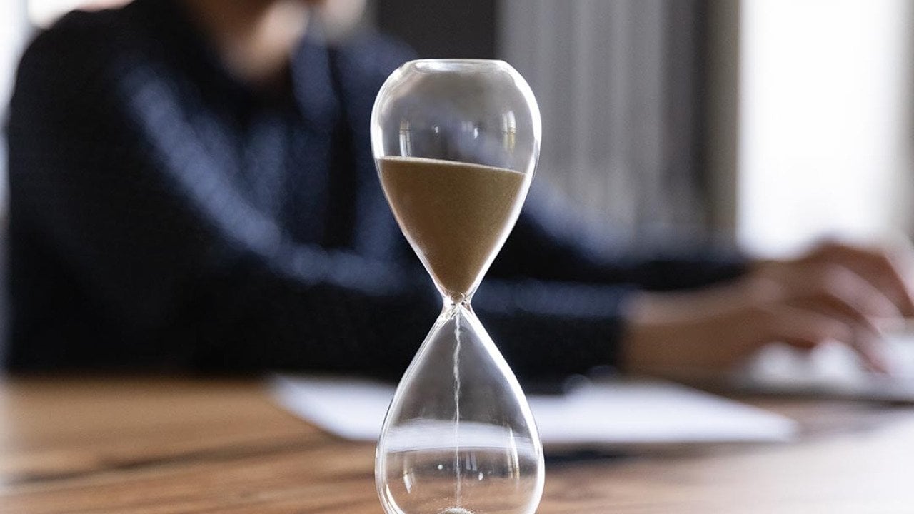 Close up view of hourglass stand on wooden office table measuring time, person working on laptop in the background blurred.