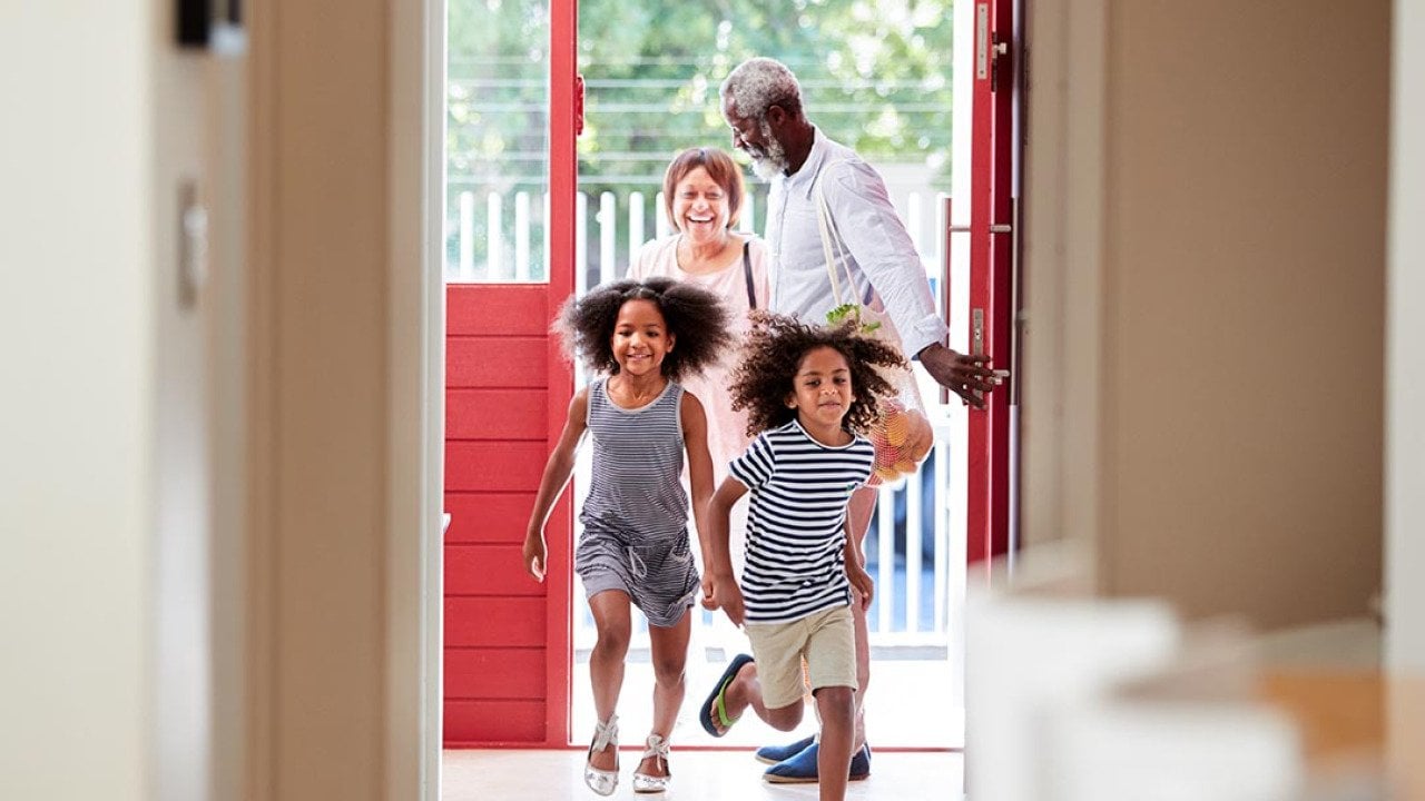 Two cute children with natural hair running into open door of house with grandparents in the background. 