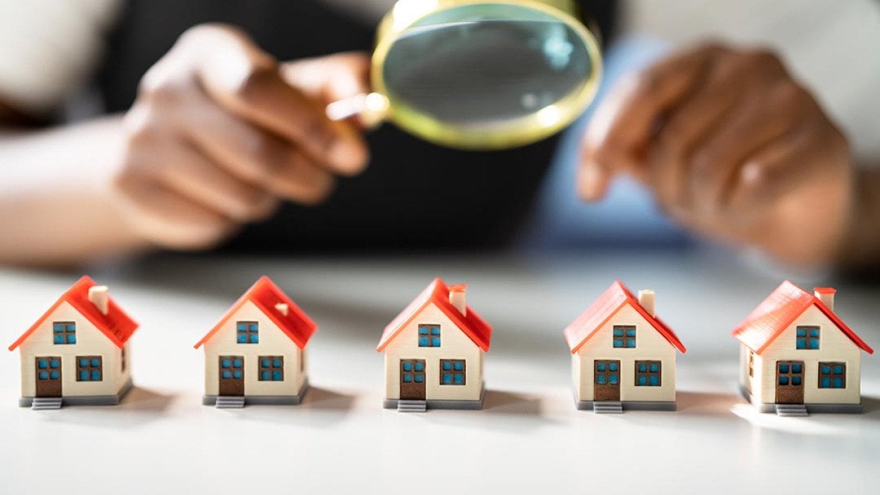 Five miniature houses with red roofs lined up on table, with magnifying glass held above them by a person symbolizing home and neighborhood appraisals.