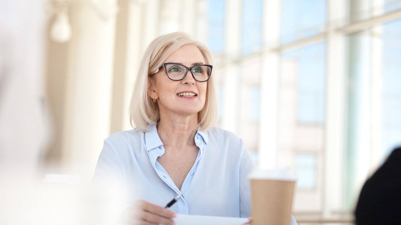 Blond professional sitting at table with large windows in the background talking in meeting.