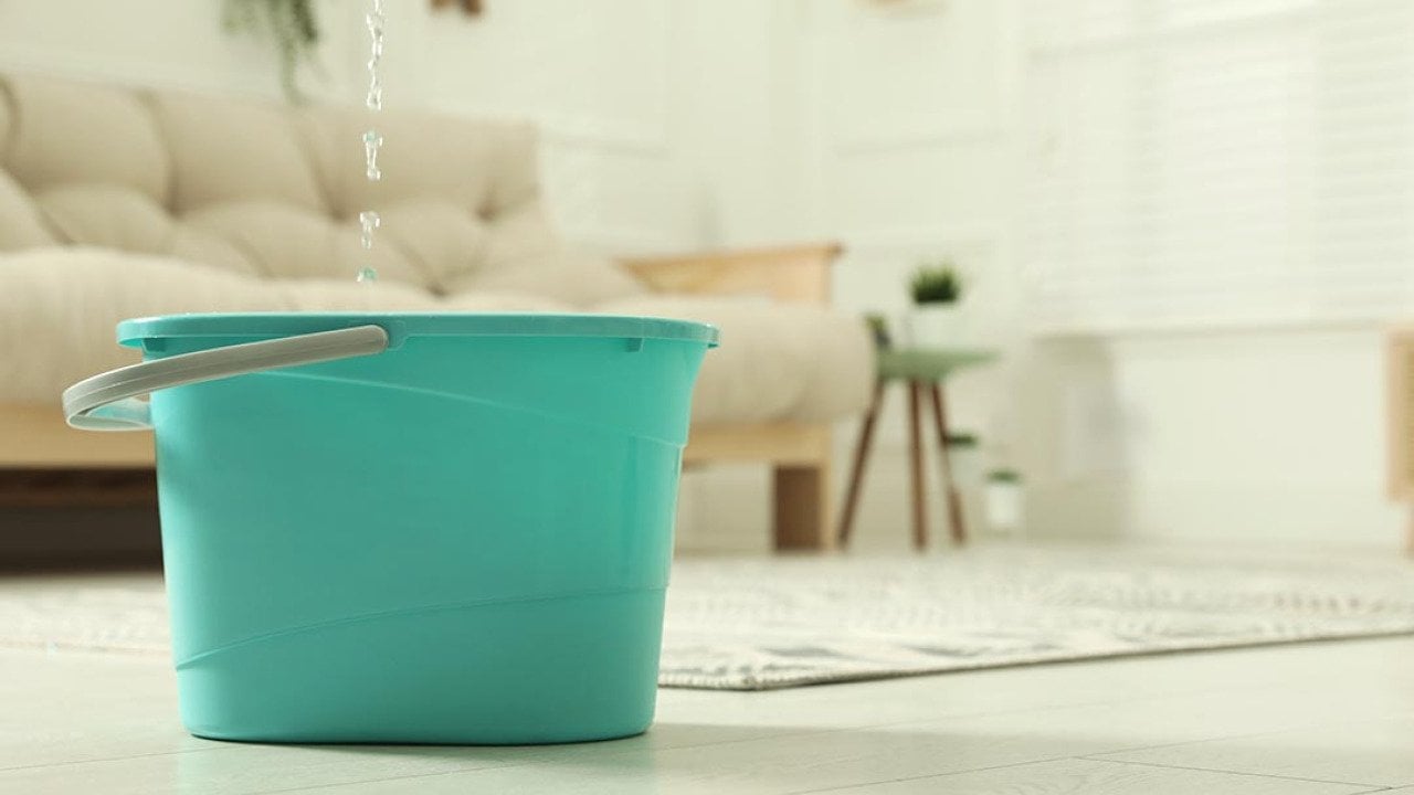 Close up of turquoise-colored plastic bucket on floor in living room with water dripping from ceiling into it.
