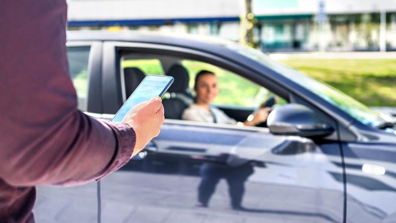 Arm of person in focus in the foreground, holding smartphone, with blurred dark car pulling up in the background, driver visible through the passenger window.
