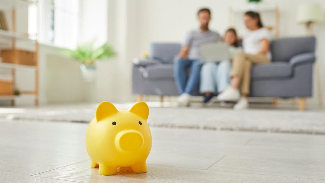 Yellow piggy money box standing on floor in the living room and a parents with a child girl sitting on sofa at home in background.