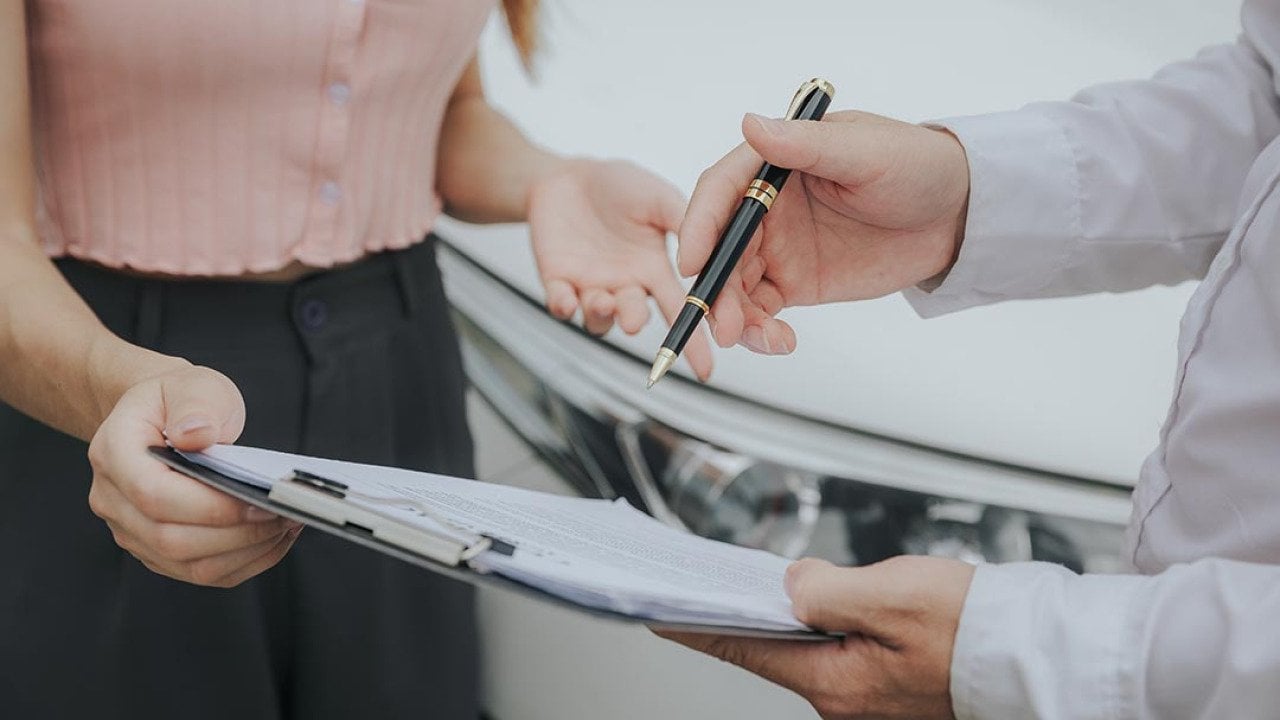 Close up of midsections of two people whose faces are not visible looking at a clipboard with forms on it with a white car in the background. 