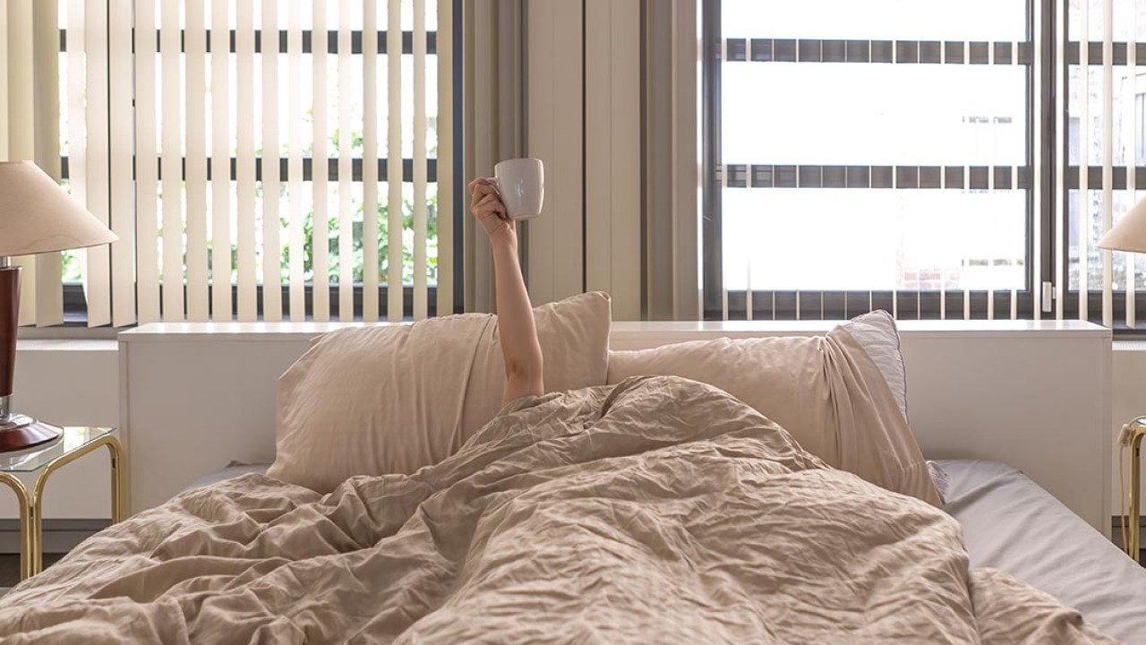 Hand holding a coffee cup extending upward from person whose face and body are under the covers in bed.
