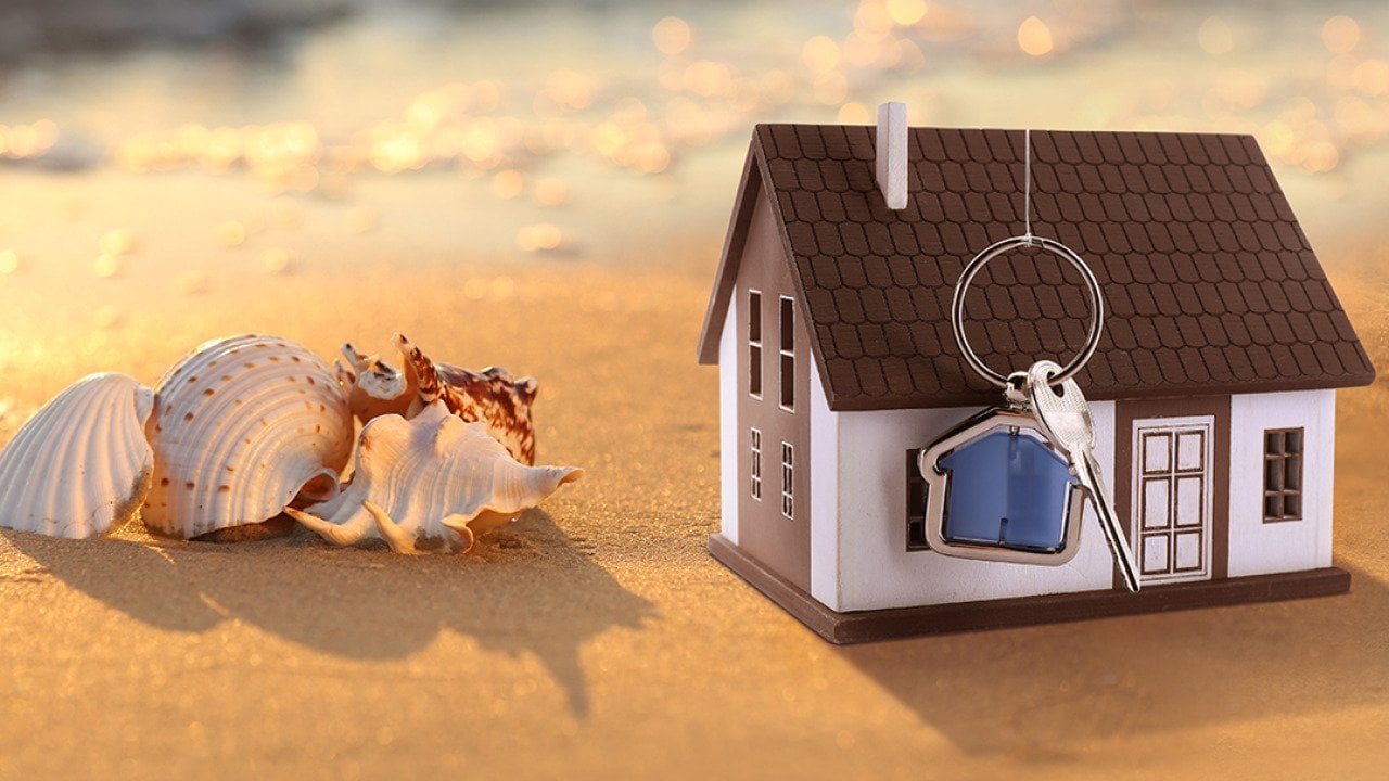 A model of a house with keys on a sandy beach beside seashells.