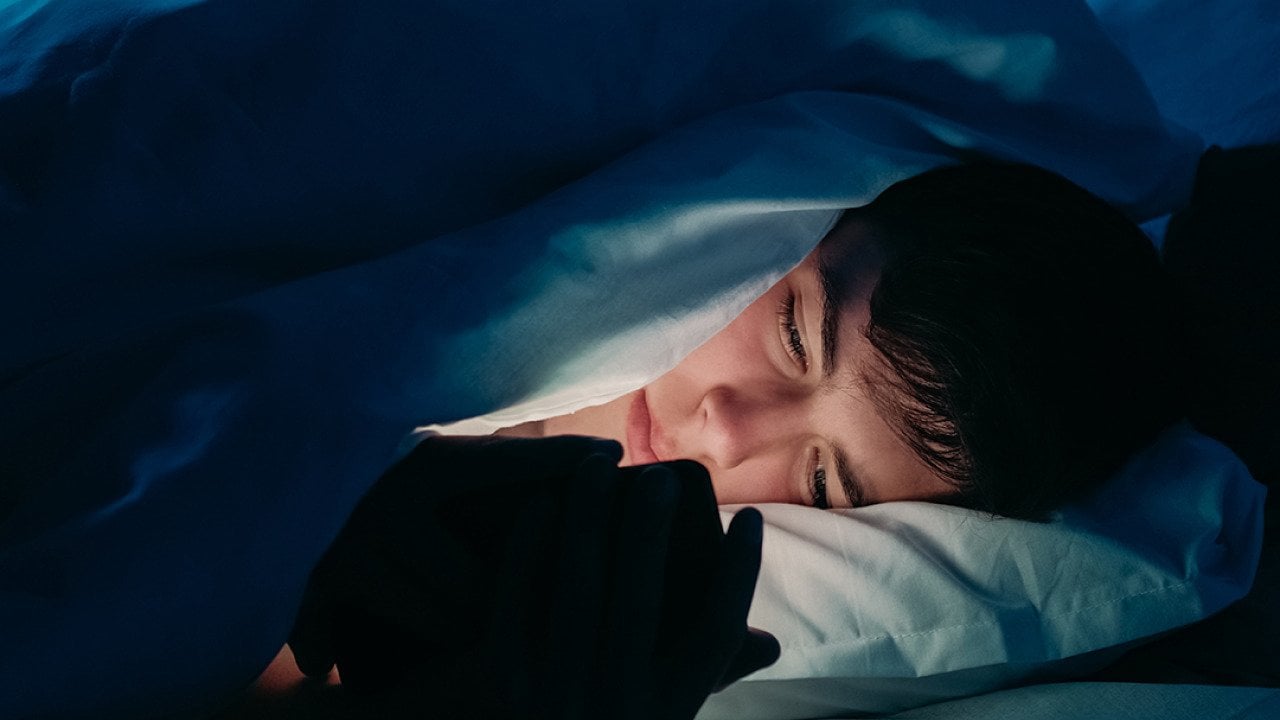 A male teenager using his smartphone under bedsheets at night.