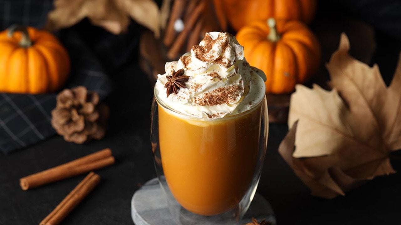 Glass of pumpkin spice latte with whipped cream and ingredients like cinnamon sticks and fall decor on black table.