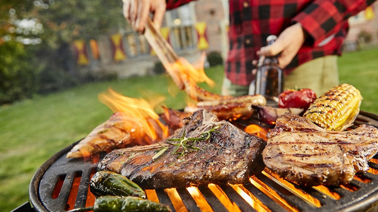 Assorted meat like sausages and steak with vegetables being grilled on a BBQ grill outdoors.