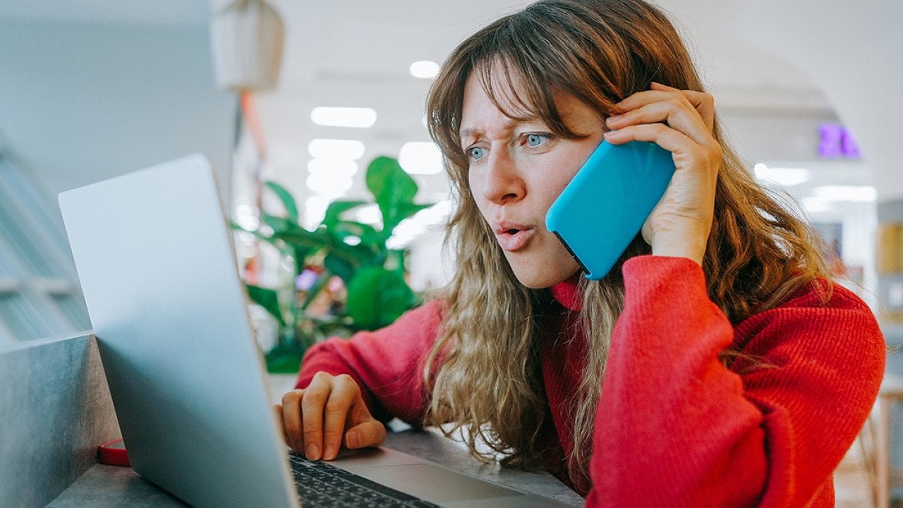 A woman talking on her smartphone while also booking flights using her laptop.